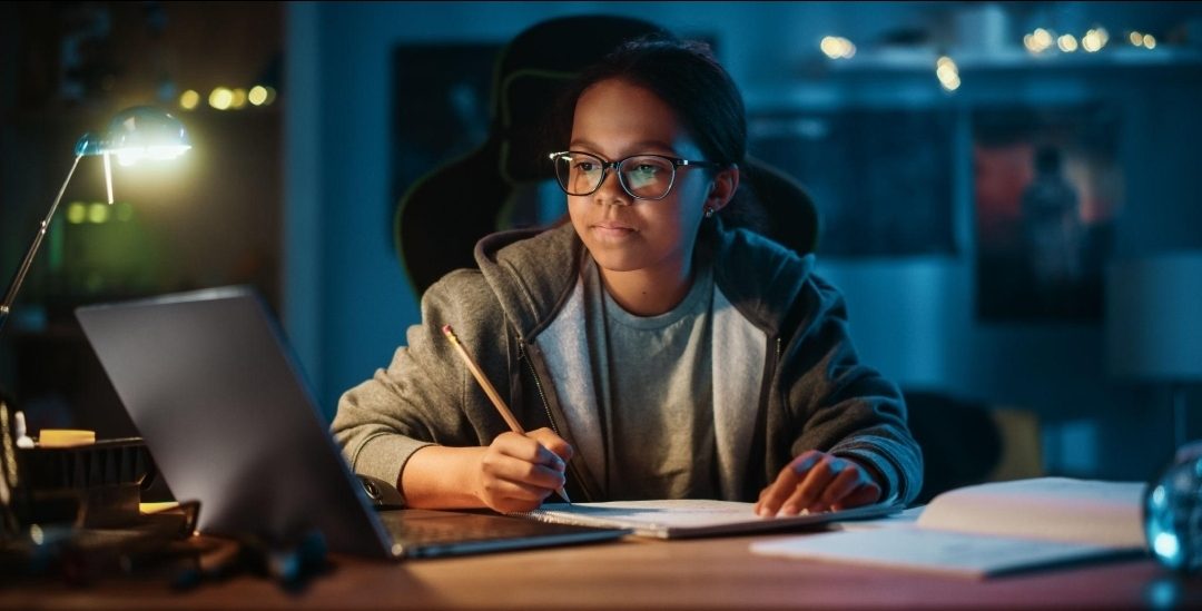 A focused young student studies at her laptop by lamplight, pencil in hand, ready to learn.